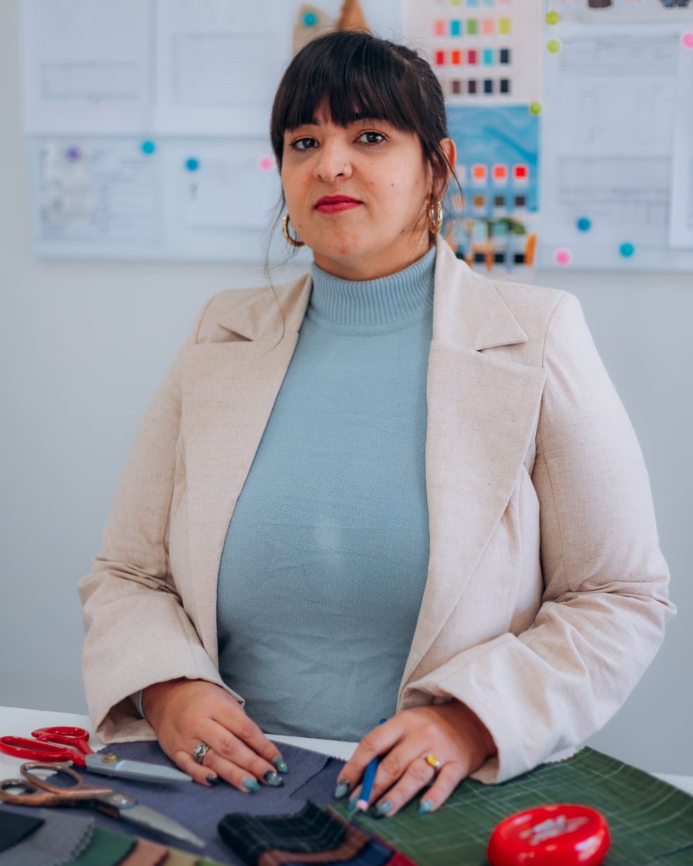 Tamara Busato à mesa de trabalho do ateliê, com amostras de tecido e o painel da coleção